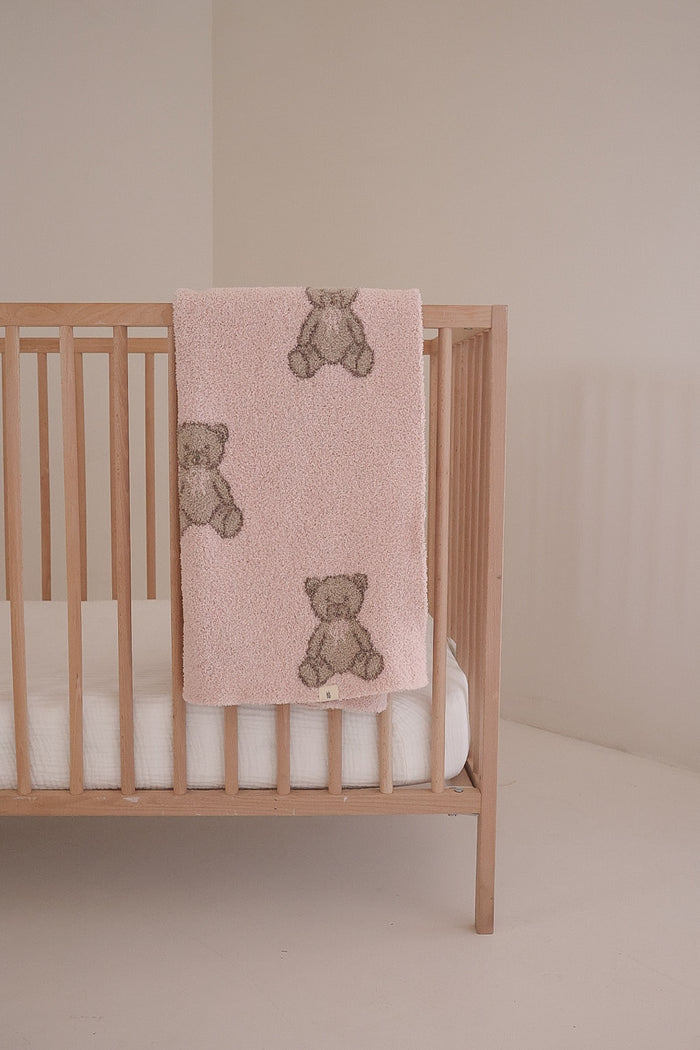 A light wooden crib with a white mattress features the forever french baby Plush Blanket | Lottie in soft pink, adorned with brown teddy bears and draped over the side, set against a plain, neutral-colored wall.