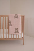 A light wooden crib with a white mattress features the forever french baby Plush Blanket | Lottie in soft pink, adorned with brown teddy bears and draped over the side, set against a plain, neutral-colored wall.
