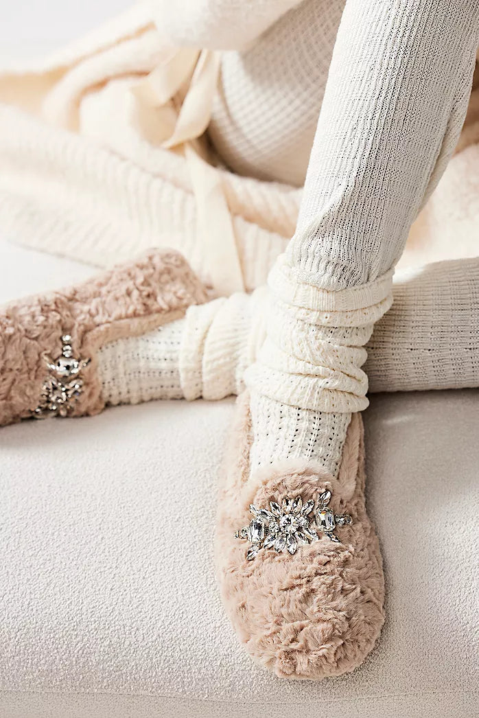 A person in cream knit loungewear and socks sits cross-legged, showcasing Free People Slumber Party Loafer slippers in blush pink with jeweled details, on a soft cream surface.