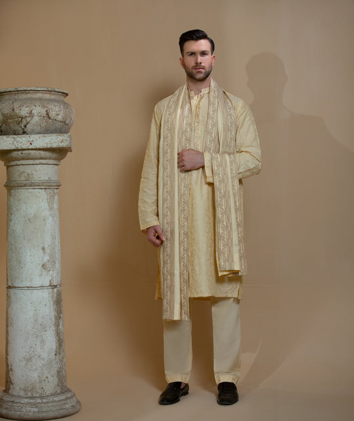 Man wearing Arsalan Iqbal's Light Gold Embroidered Sherwani Coat over a matching kurta pyjama, with a gold-cream stole draped over shoulders and dark formal shoes, posed beside a stone pillar on a beige backdrop