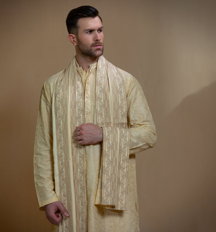 Man wearing Arsalan Iqbal's Light Gold Embroidered Sherwani Coat over a matching kurta pyjama, with a gold-cream stole draped over shoulders and dark formal shoes, posed beside a stone pillar on a beige backdrop