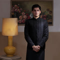 Arsalan Iqbal Jet Black Hand Embroidered Prince Jacket with Kurta Pyjama man wearing a fully embellished jet black prince jacket featuring dense hand embroidery with sequin and thread detailing, paired with a matching black kurta pyjama, mandarin collar, and a sleek formal silhouette, styled in a warm indoor setting
