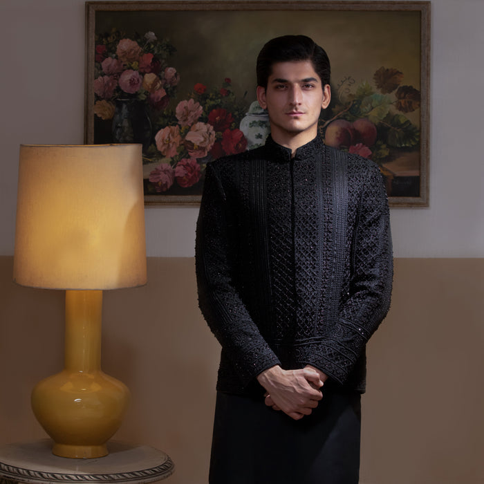 Arsalan Iqbal Jet Black Hand Embroidered Prince Jacket with Kurta Pyjama man wearing a fully embellished jet black prince jacket featuring dense hand embroidery with sequin and thread detailing, paired with a matching black kurta pyjama, mandarin collar, and a sleek formal silhouette, styled in a warm indoor setting