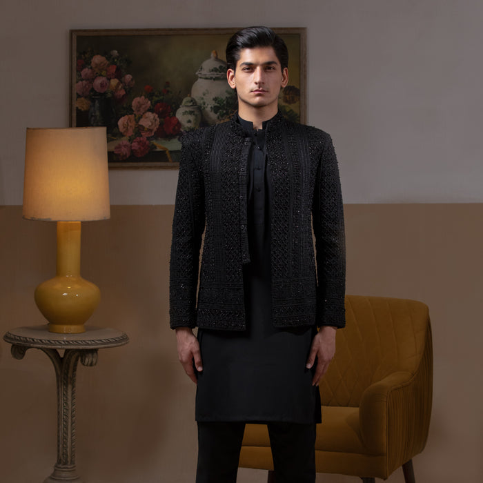 Arsalan Iqbal Jet Black Hand Embroidered Prince Jacket with Kurta Pyjama man wearing a fully embellished jet black prince jacket featuring dense hand embroidery with sequin and thread detailing, paired with a matching black kurta pyjama, mandarin collar, and a sleek formal silhouette, styled in a warm indoor setting