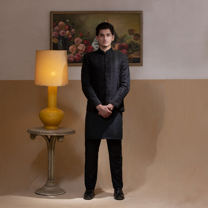 Arsalan Iqbal Jet Black Hand Embroidered Prince Jacket with Kurta Pyjama man wearing a fully embellished jet black prince jacket featuring dense hand embroidery with sequin and thread detailing, paired with a matching black kurta pyjama, mandarin collar, and a sleek formal silhouette, styled in a warm indoor setting