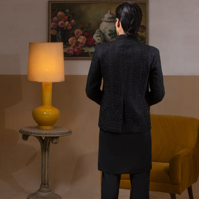 Arsalan Iqbal Jet Black Hand Embroidered Prince Jacket with Kurta Pyjama  man wearing a fully embellished jet black prince jacket featuring dense hand embroidery with sequin and thread detailing, paired with a matching black kurta pyjama, mandarin collar, and a sleek formal silhouette, styled in a warm indoor setting