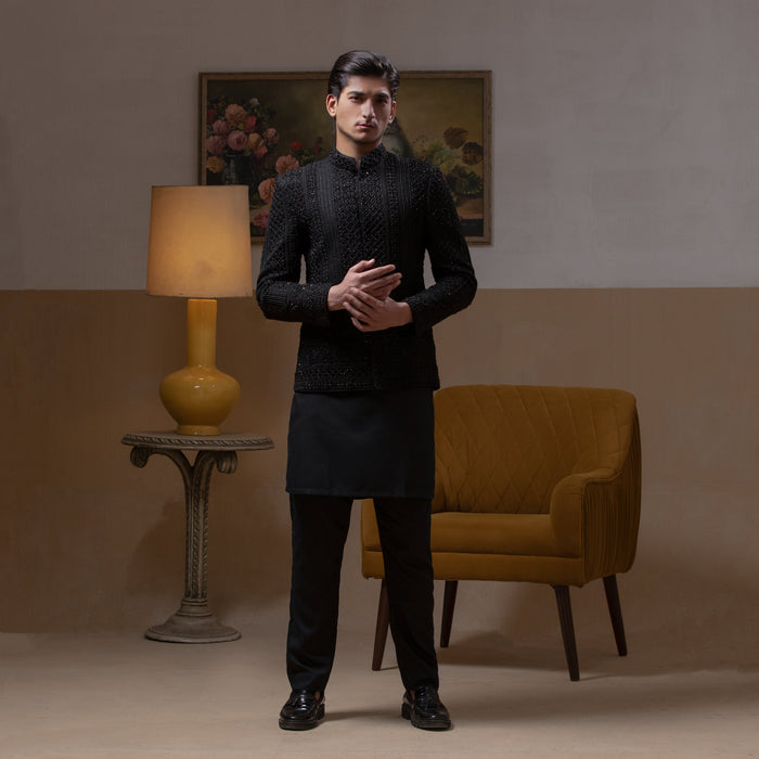 Arsalan Iqbal Jet Black Hand Embroidered Prince Jacket with Kurta Pyjama  man wearing a fully embellished jet black prince jacket featuring dense hand embroidery with sequin and thread detailing, paired with a matching black kurta pyjama, mandarin collar, and a sleek formal silhouette, styled in a warm indoor setting