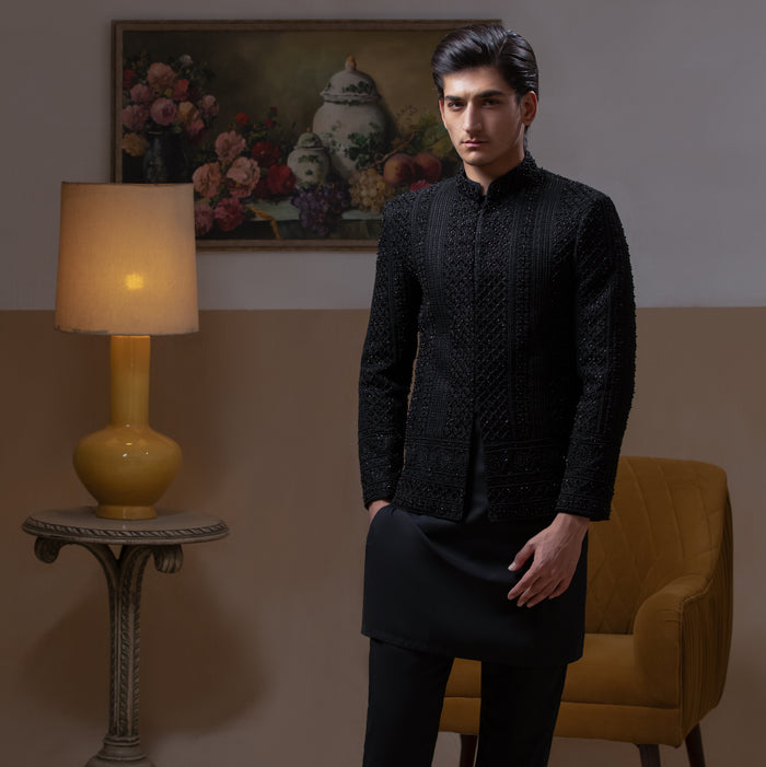 Arsalan Iqbal Jet Black Hand Embroidered Prince Jacket with Kurta Pyjama – man wearing a fully embellished jet black prince jacket featuring dense hand embroidery with sequin and thread detailing, paired with a matching black kurta pyjama, mandarin collar, and a sleek formal silhouette, styled in a warm indoor setting