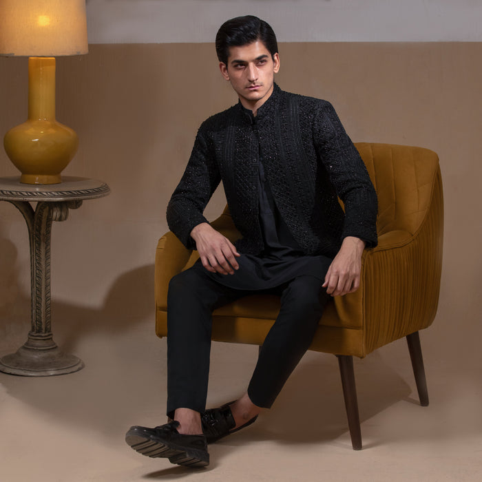 Arsalan Iqbal Jet Black Hand Embroidered Prince Jacket with Kurta Pyjama man wearing a fully embellished jet black prince jacket featuring dense hand embroidery with sequin and thread detailing, paired with a matching black kurta pyjama, mandarin collar, and a sleek formal silhouette, styled in a warm indoor setting