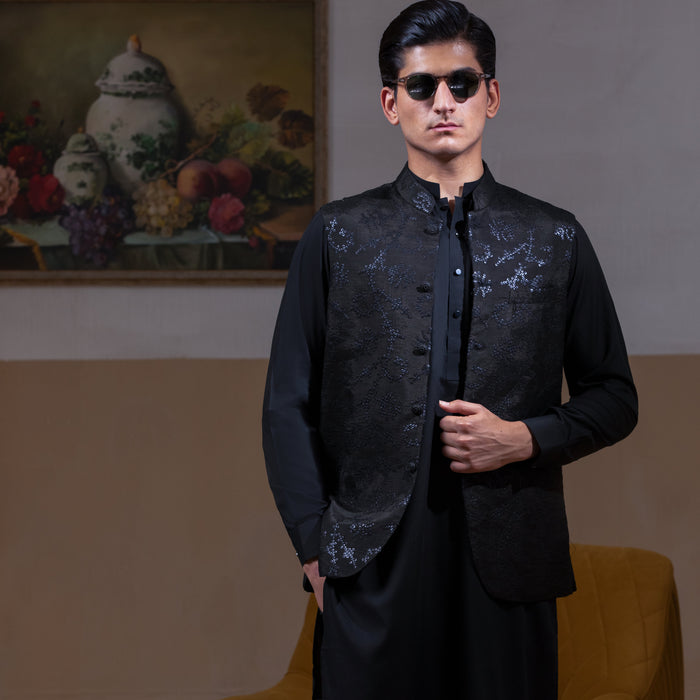 Man wearing Arsalan Iqbal's Black Sequin Embroidered Waistcoat over a matching black kurta pyjama, styled with embellished loafers and sunglasses, posed beside a mustard chair against a beige backdrop