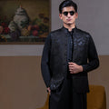 Man wearing Arsalan Iqbal's Black Sequin Embroidered Waistcoat over a matching black kurta pyjama, styled with embellished loafers and sunglasses, posed beside a mustard chair against a beige backdrop