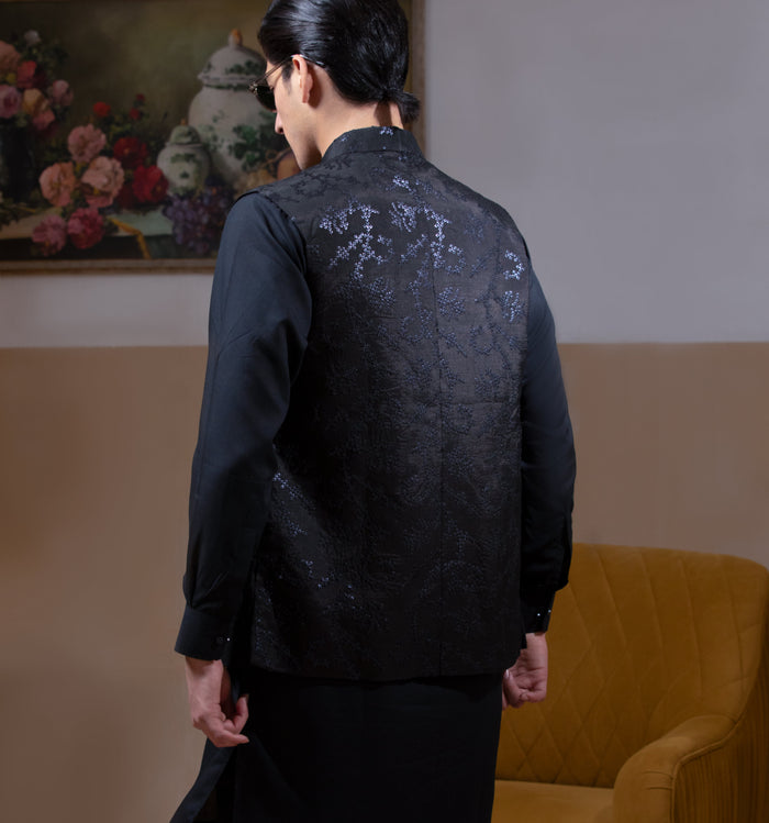 Man wearing Arsalan Iqbal's Black Sequin Embroidered Waistcoat over a matching black kurta pyjama, styled with embellished loafers and sunglasses, posed beside a mustard chair against a beige backdrop