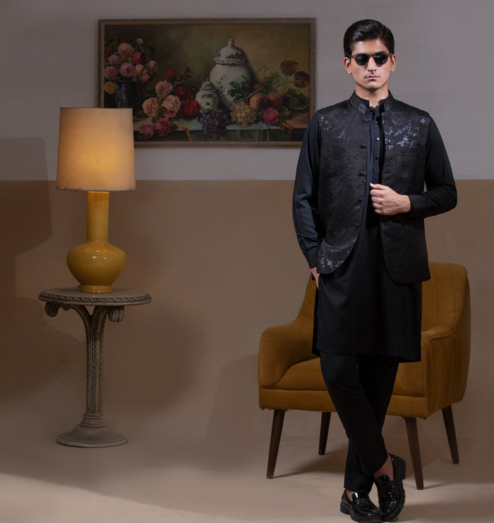 Man wearing Arsalan Iqbal's Black Sequin Embroidered Waistcoat over a matching black kurta pyjama, styled with embellished loafers and sunglasses, posed beside a mustard chair against a beige backdrop