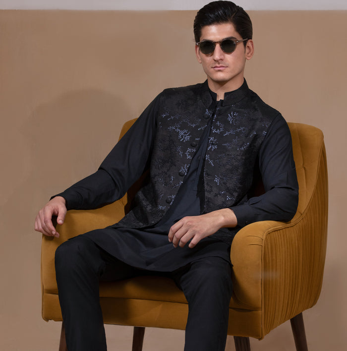 Man wearing Arsalan Iqbal's Black Sequin Embroidered Waistcoat over a matching black kurta pyjama, styled with embellished loafers and sunglasses, posed beside a mustard chair against a beige backdrop