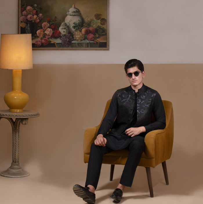 Man wearing Arsalan Iqbal's Black Sequin Embroidered Waistcoat over a matching black kurta pyjama, styled with embellished loafers and sunglasses, posed beside a mustard chair against a beige backdrop