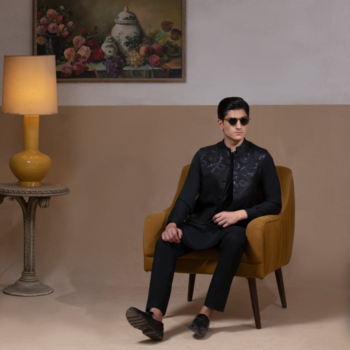 Man wearing Arsalan Iqbal's Black Sequin Embroidered Waistcoat over a matching black kurta pyjama, styled with embellished loafers and sunglasses, posed beside a mustard chair against a beige backdrop