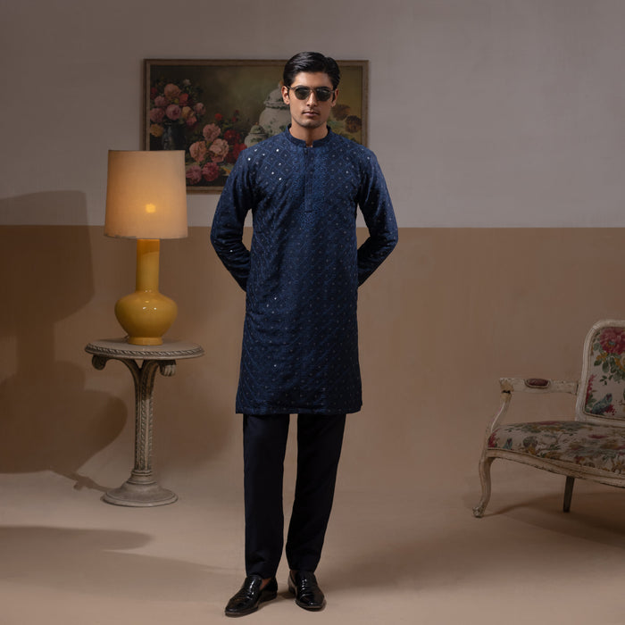 Man wearing Arsalan Iqbal's Navy Blue Allover Sequin Embroidered Kurta with dark navy trousers, black loafers and sunglasses, posed against a beige backdrop with yellow lamp and floral painting