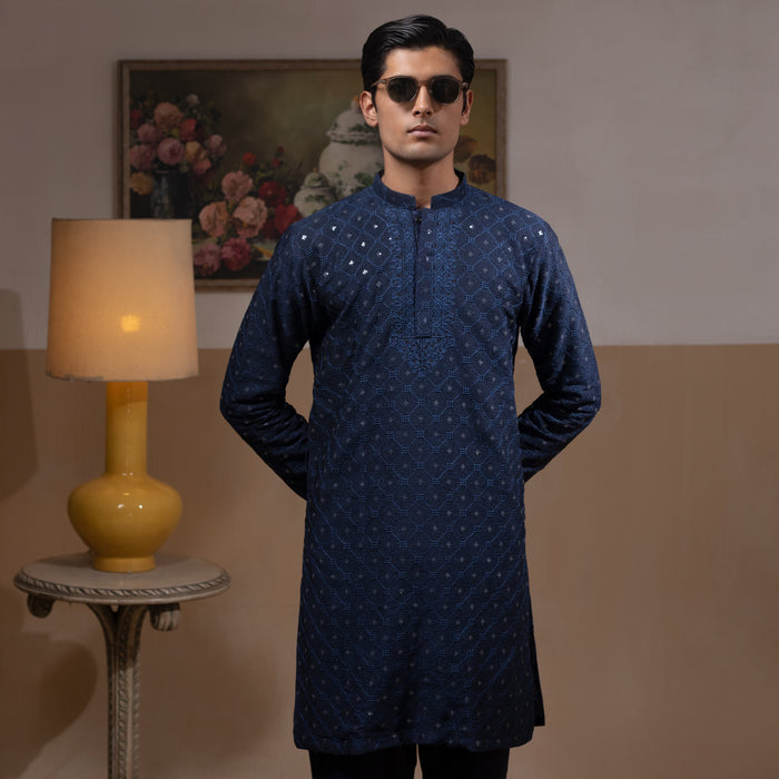 Man wearing Arsalan Iqbal's Navy Blue Allover Sequin Embroidered Kurta with dark navy trousers, black loafers and sunglasses, posed against a beige backdrop with yellow lamp and floral painting