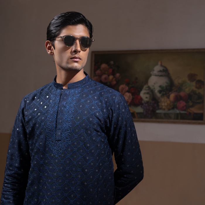 Man wearing Arsalan Iqbal's Navy Blue Allover Sequin Embroidered Kurta with dark navy trousers, black loafers and sunglasses, posed against a beige backdrop with yellow lamp and floral painting
