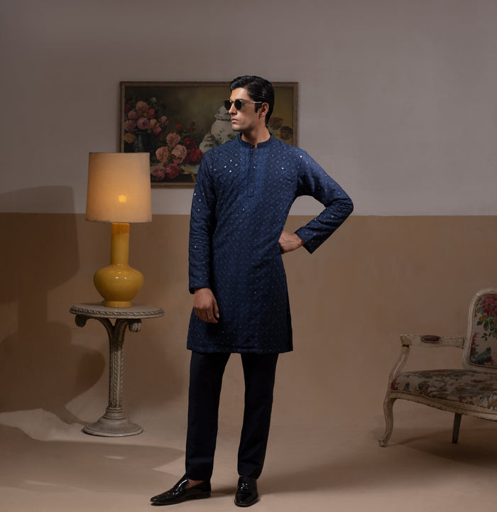 Man wearing Arsalan Iqbal's Navy Blue Allover Sequin Embroidered Kurta with dark navy trousers, black loafers and sunglasses, posed against a beige backdrop with yellow lamp and floral painting