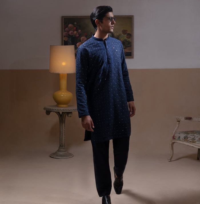 Man wearing Arsalan Iqbal's Navy Blue Allover Sequin Embroidered Kurta with dark navy trousers, black loafers and sunglasses, posed against a beige backdrop with yellow lamp and floral painting