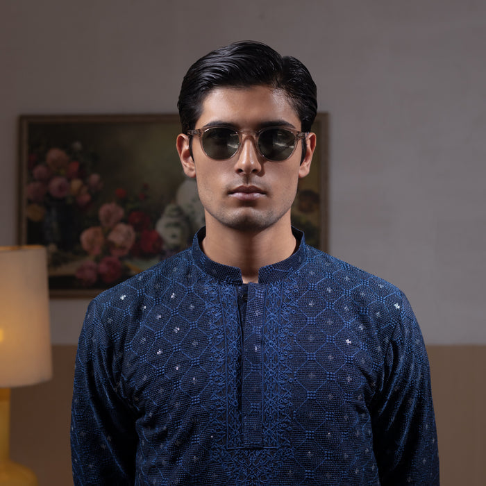 Man wearing Arsalan Iqbal's Navy Blue Allover Sequin Embroidered Kurta with dark navy trousers, black loafers and sunglasses, posed against a beige backdrop with yellow lamp and floral painting