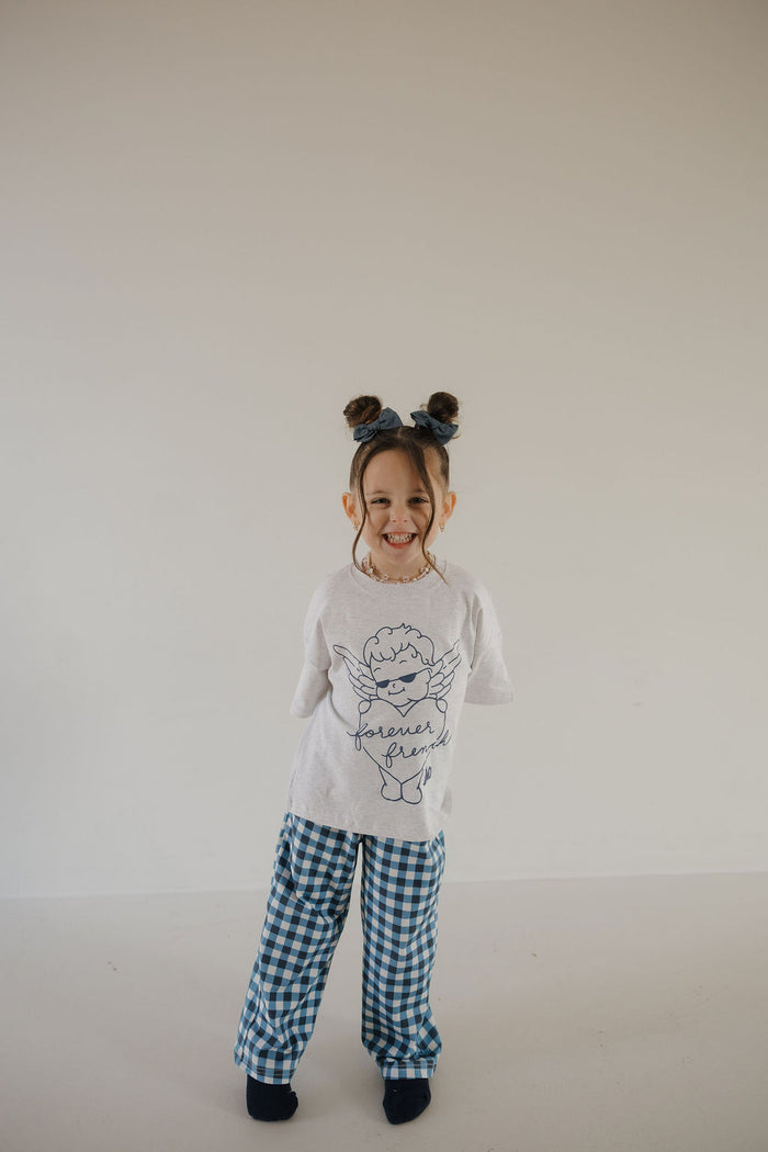 A young child with brown hair in two buns models the forever french baby T-Shirt & Pant Set from ff Los Angeles, featuring a white tee with angel graphic and "forever young" text plus blue gingham pants—perfect for trendy LA kids.
