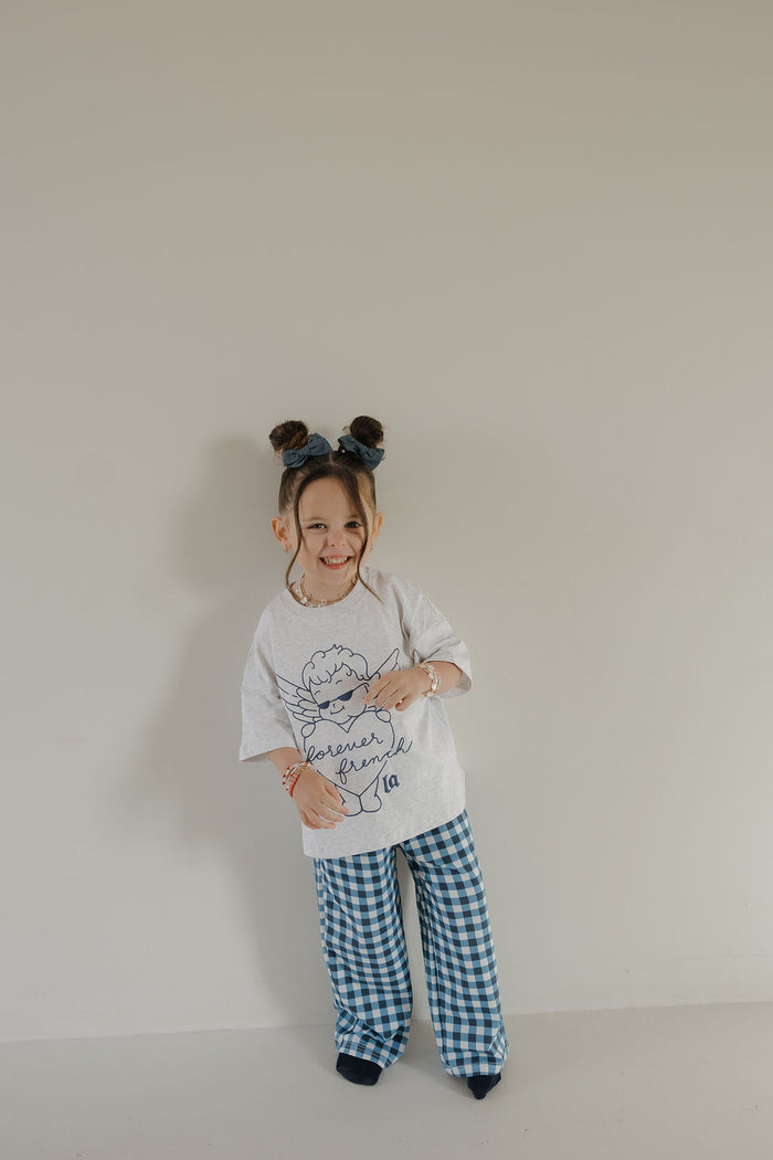 A young child with two hair buns and a blue headband smiles in the T-Shirt & Pant Set by forever french baby, standing against a light wall—perfect stylish kids clothing from ff Los Angeles.
