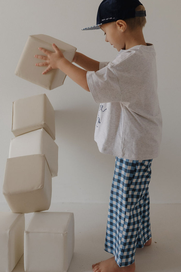A young child in the forever french baby T-Shirt & Pant Set by ff Los Angeles, wearing a cap and blue gingham pants, carefully stacks large white foam blocks as the tower begins to wobble.