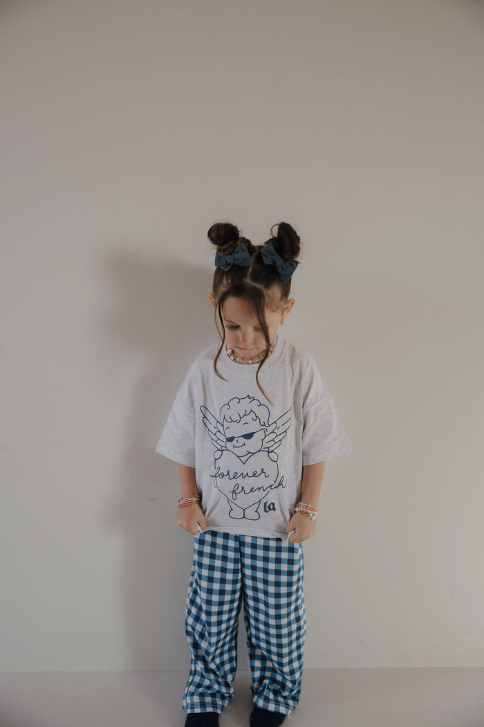 A young child with two hair buns and blue bows stands against a plain wall, looking down. They wear the forever french baby T-Shirt & Pant Set from ff Los Angeles: a white tee with a cartoon angel and blue lettering, plus gingham pants.