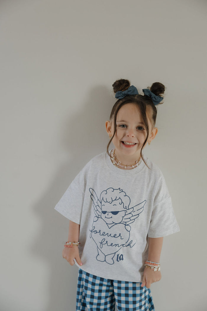 A young child smiles at the camera in the forever french baby T-Shirt & Pant Set | ff Los Angeles, featuring an angel graphic top and blue gingham pants, styled with a navy hair bow and beaded jewelry—perfect for kids in LA.