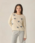 A young person wears the Rylee + Cru Teen Crochet Knit Pullover | Bows—featuring a cream crochet design with blue bow details—and striped pants, standing against a plain light background while looking at the camera with a neutral expression.