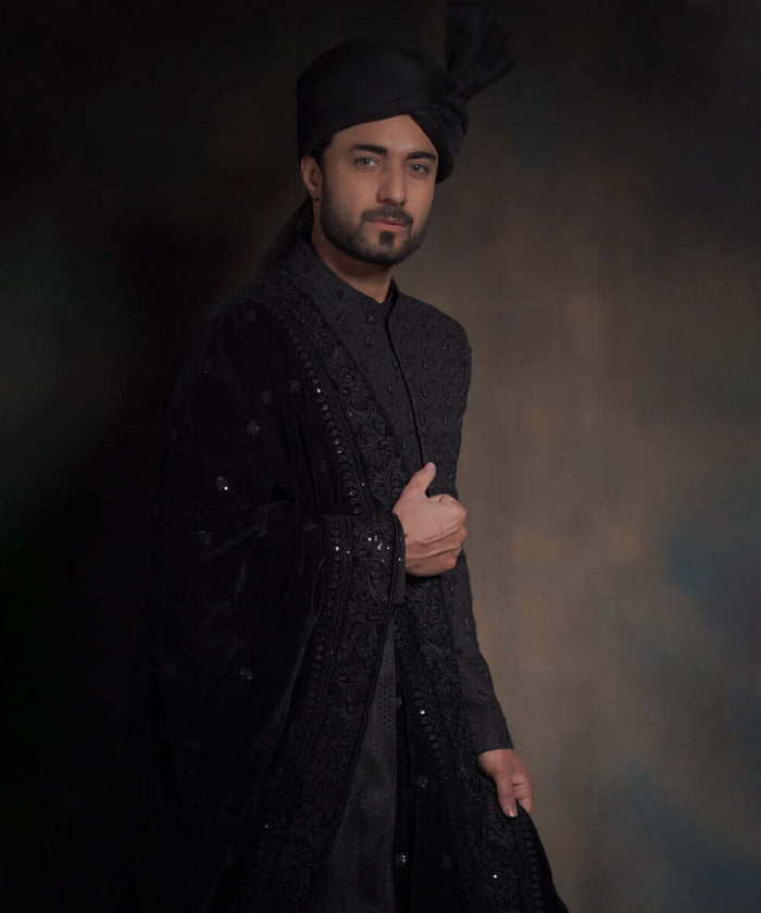 Portrait of Arsalan Iqbal midnight black sherwani with spaced-out tonal floral motifs, featuring a draped velvet shawl with heavy embroidery and a matching high-pleated turban.