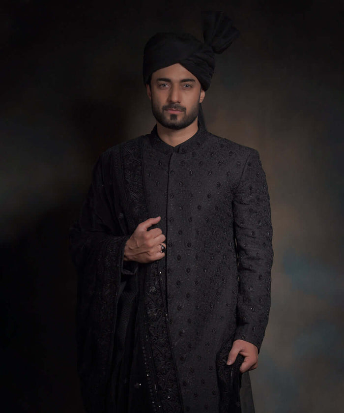 Front portrait of Arsalan Iqbal midnight black luxury sherwani featuring spaced-out tonal floral motifs, paired with a matching black turban and a heavily embroidered draped shawl.