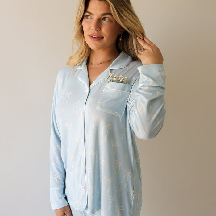 A woman with blonde hair wears the forever french baby Women's Bamboo Button Up Lounge Set | Blue Bows, standing against a plain background, touching her hair with white flowers tucked into her chest pocket.