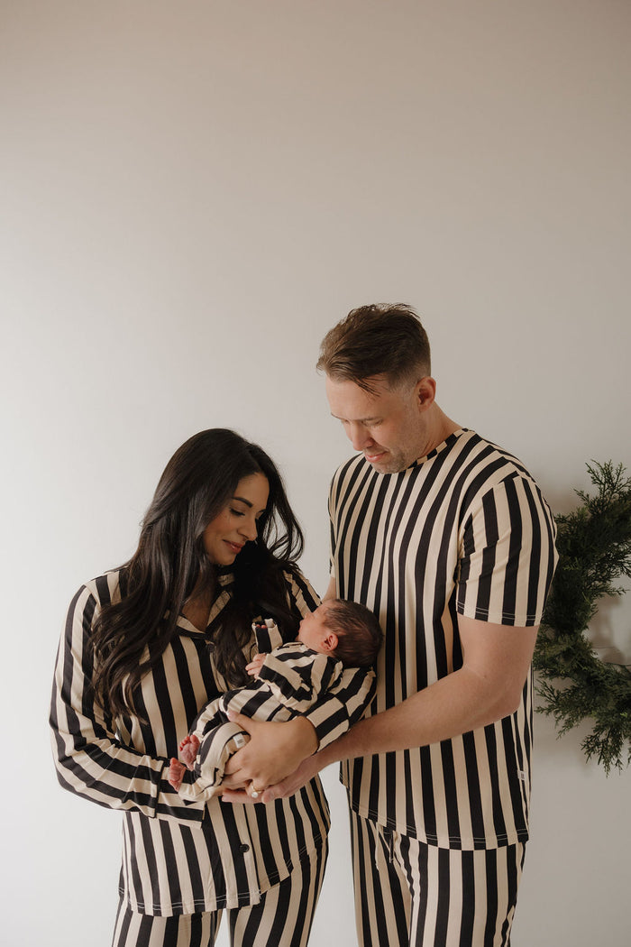 A man and woman, both wearing forever french baby's Midnight Stripe bamboo pajamas, lovingly hold a baby in matching sleepwear. They stand before a white wall with a green wreath partly visible on the right.