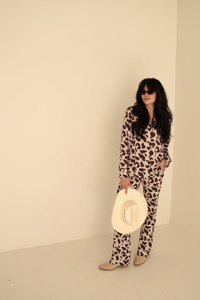 A woman models the forever french baby Women's Bamboo Button Up Lounge Set | Moo! in a cow-print pattern, paired with a white cowboy hat and beige shoes—perfect inspiration for styling hypoallergenic fashion confidently.