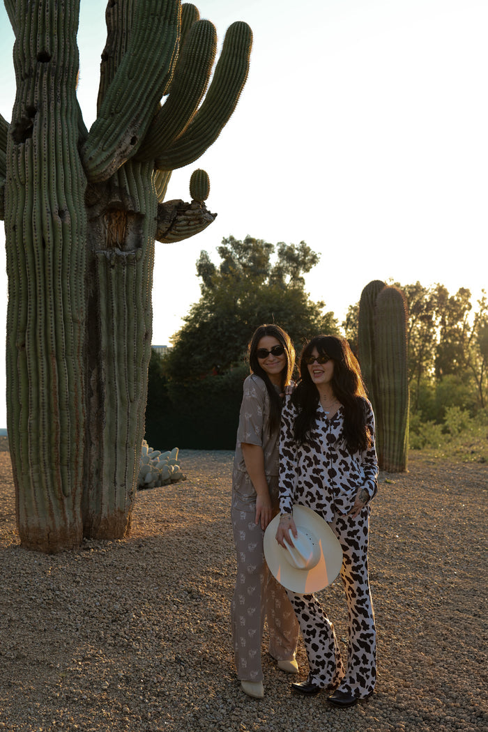 Two women wearing the Women's Bamboo Button Up Lounge Set by forever french baby smile near desert cacti at sunset. One holds a white hat; both sport sunglasses and long dark hair. Trees can be seen in the background.