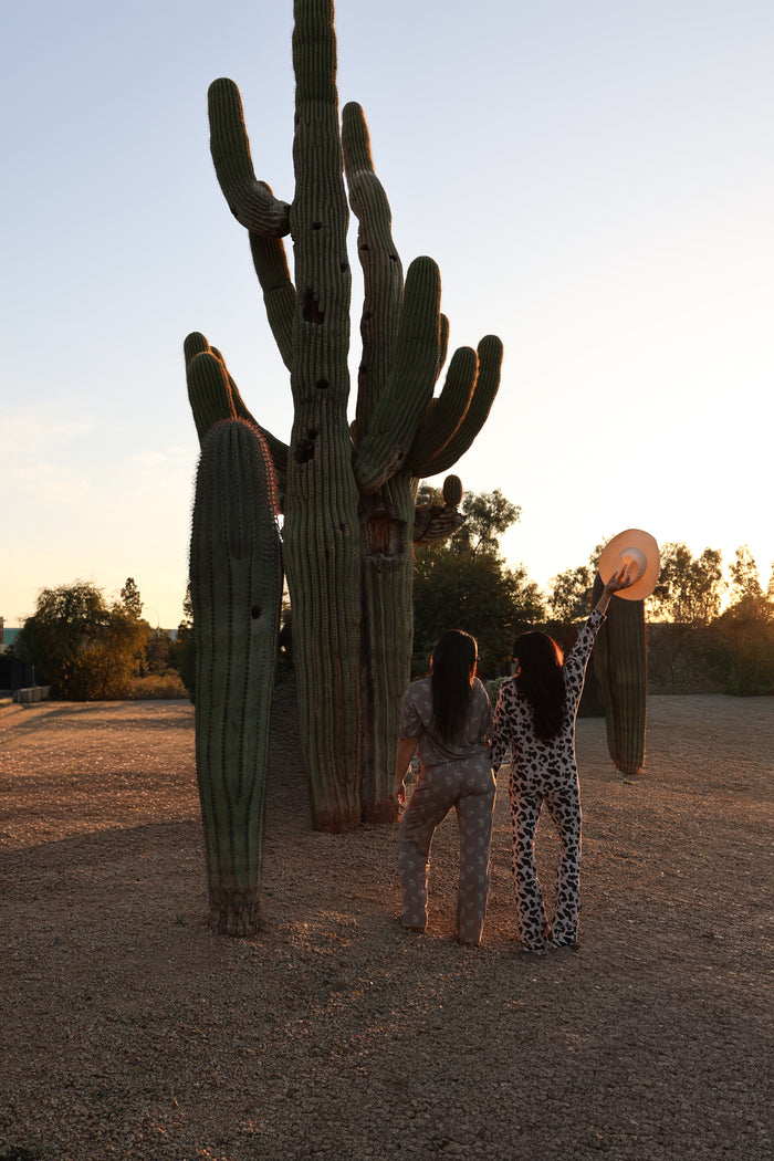 Two people wear the Women's Bamboo Button Up Lounge Set by forever french baby, standing hand in hand near tall cacti at sunset; one lifts a hat as warm sunlight casts long shadows across the desert ground.
