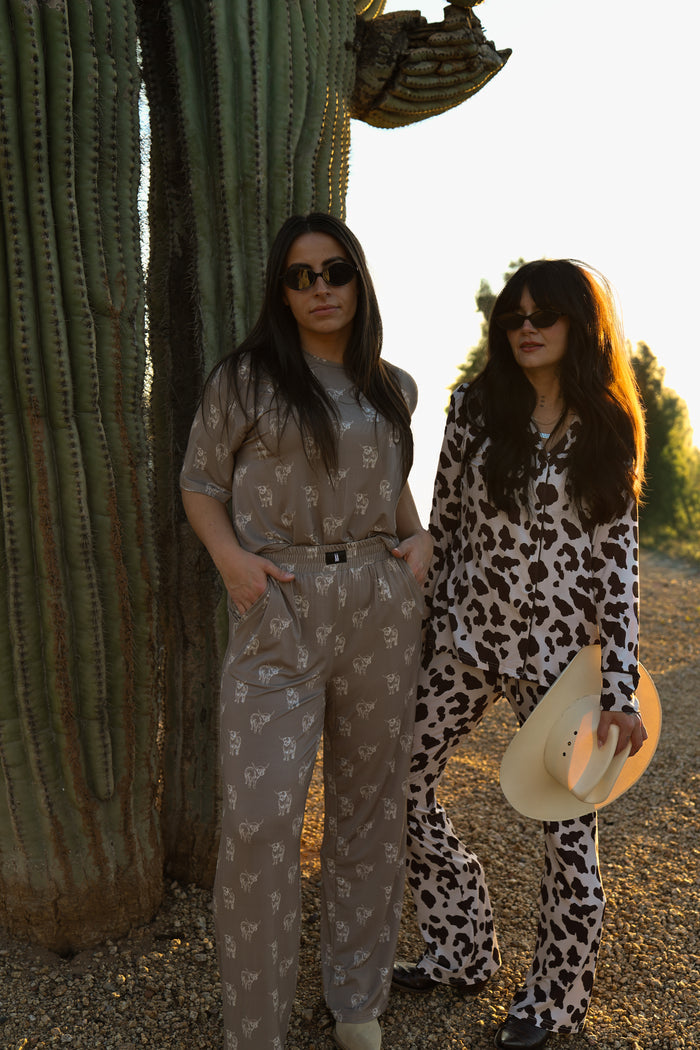 Two women in forever french baby's Women's Boxy Tee Lounge Set | Highland pose by a cactus. One wears grey with white prints, the other rocks a black-and-white cow print set, both sporting sunglasses and basking in the sun in comfy bamboo loungewear.