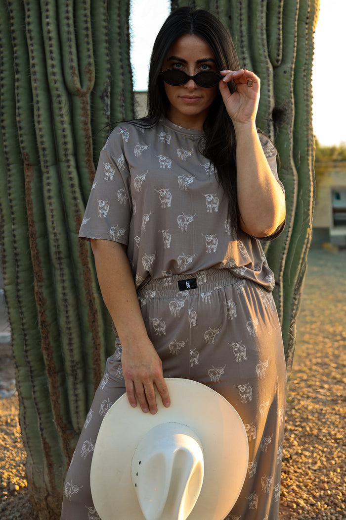 A woman wears the forever french baby Women's Boxy Tee Lounge Set in Highland, standing by a cactus with a white cowboy hat in hand and adjusting her dark sunglasses.