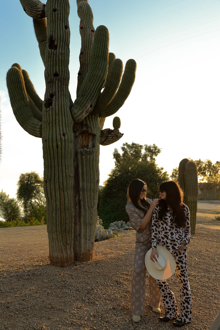 Two women in Women's Bamboo Button Up Lounge Sets by forever french baby stand by a cactus in a sunny desert. One adjusts the other's hair while holding a hat, surrounded by cacti and trees, showcasing their hypoallergenic outfits.