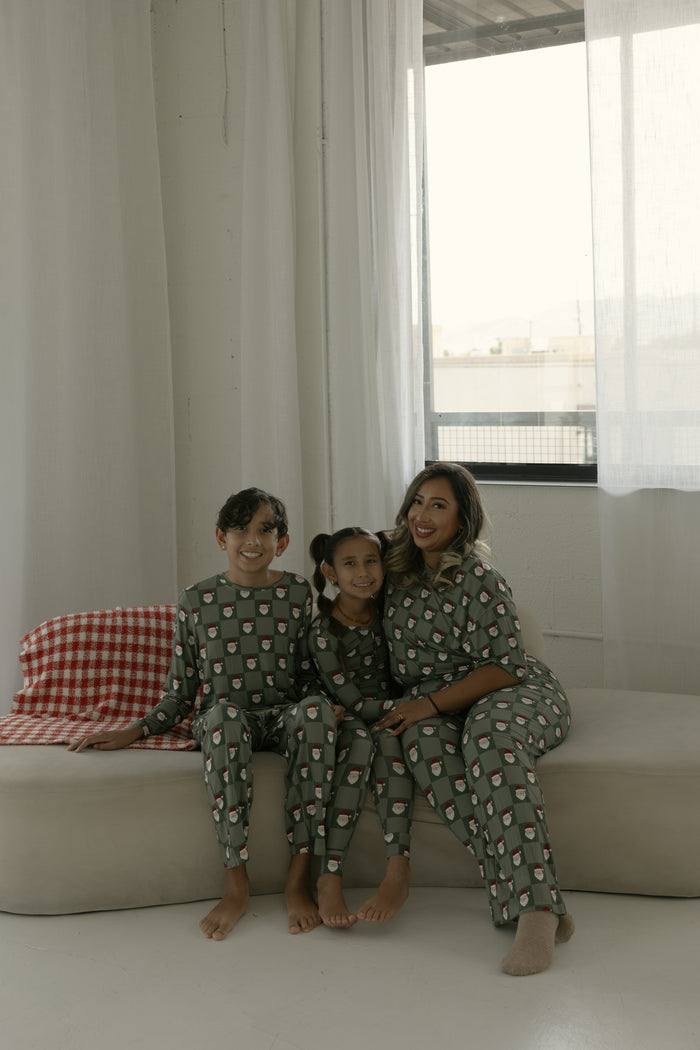 A woman and two children smile on a beige couch, wearing matching forever french baby Pre-Teen Straight Leg Bamboo Pajamas in the Santa Checkerboard pattern. Sheer white curtains frame the cozy scene in the background.