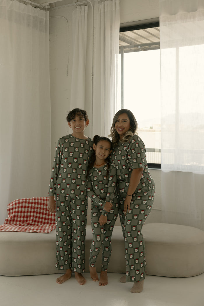 A woman and two children smile indoors, barefoot in matching forever french baby Pre-Teen Straight Leg Bamboo Pajamas | Santa Checkerboard, posing by sheer white curtains and a checkered blanket on a bench.