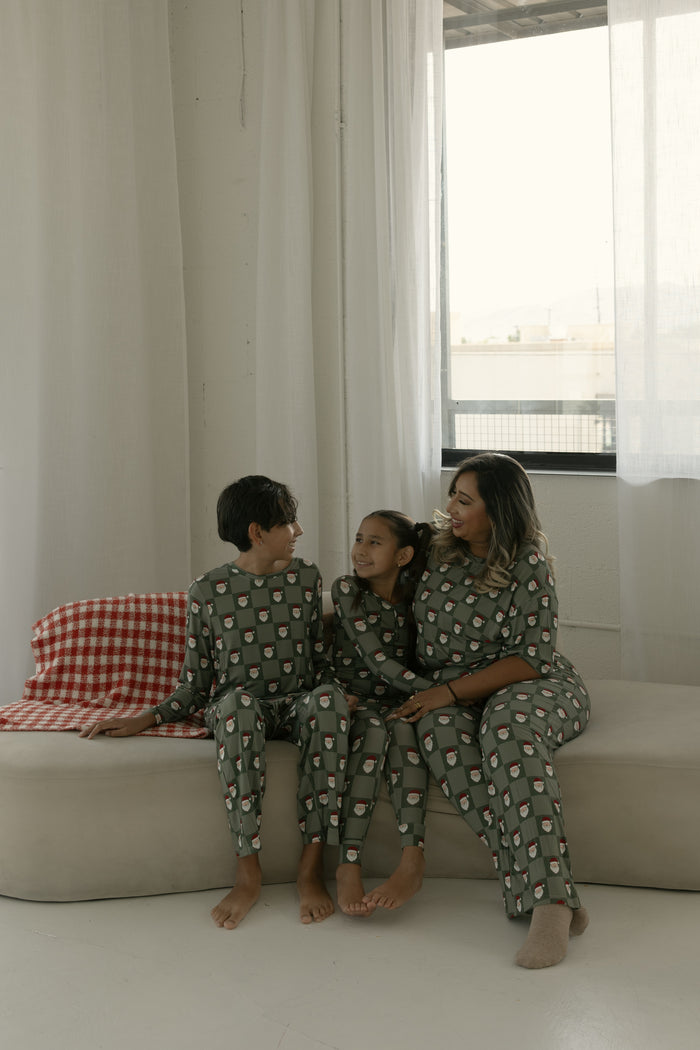 A woman and two children in forever french baby Pre-Teen Straight Leg Bamboo Pajamas | Santa Checkerboard sit smiling on a beige couch with white curtains and a red checked blanket visible in the room.